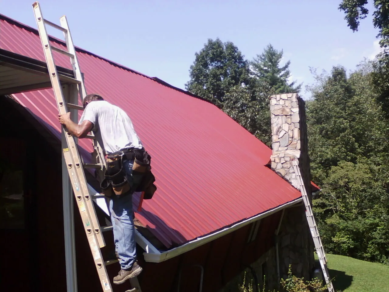 Roofer on a ladder beside a red metal roof and stone chimney — Soffit & Fascia Repair by Roofing Brothers in Beaman